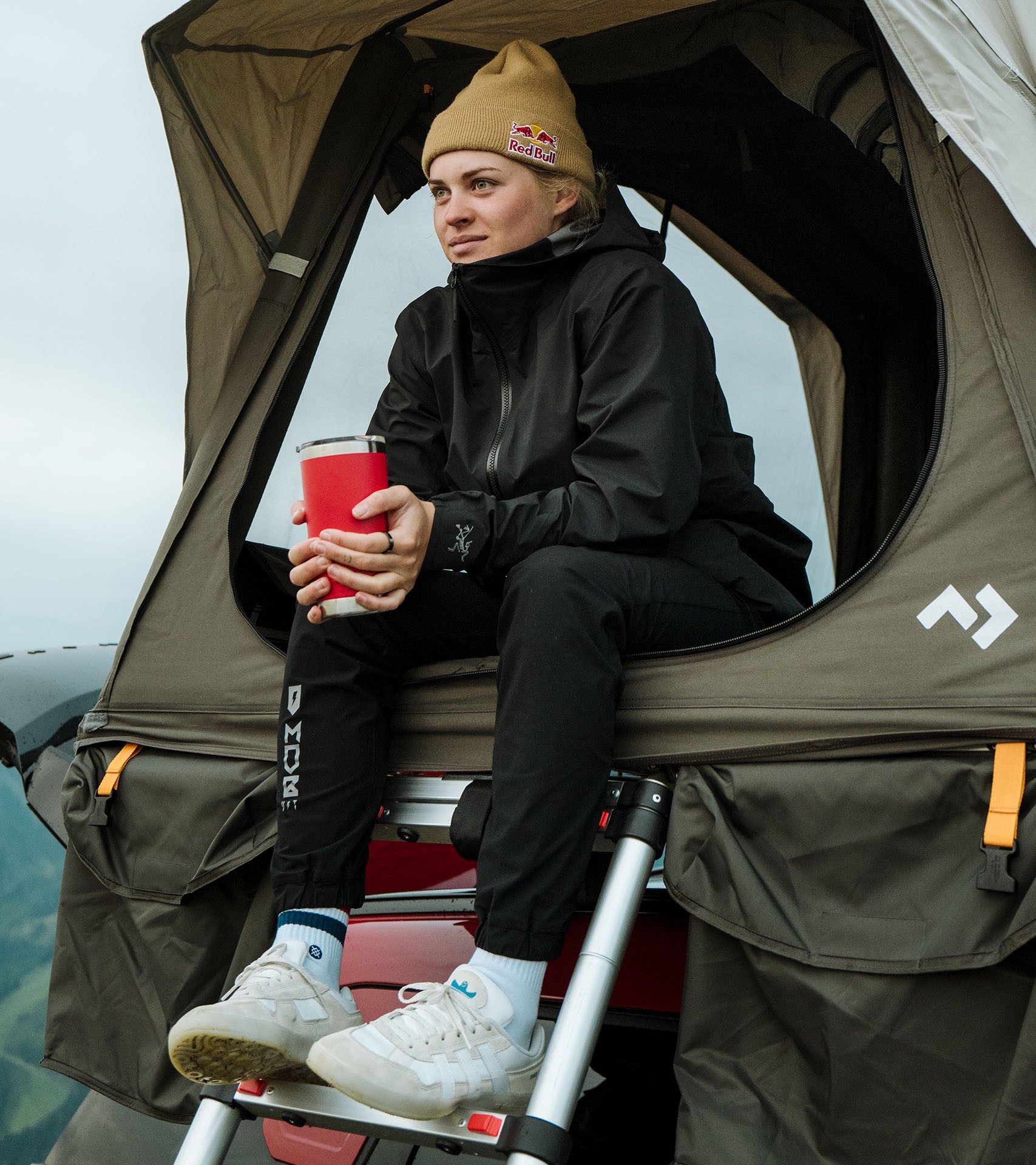 In warm clothing and a Redbull beanie, Vali holds a red cup and sits atop the step ladder leading into a roof tent on the new MINI John Cooper Works Countryman.